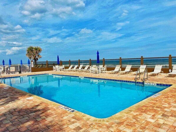 A pool with a clear blue sky and ocean view, surrounded by lounge chairs and umbrellas. - NEW Amazing Spacious Ocean View Studio in Daytona - Up 6 Guests! (Daytona Beach)
