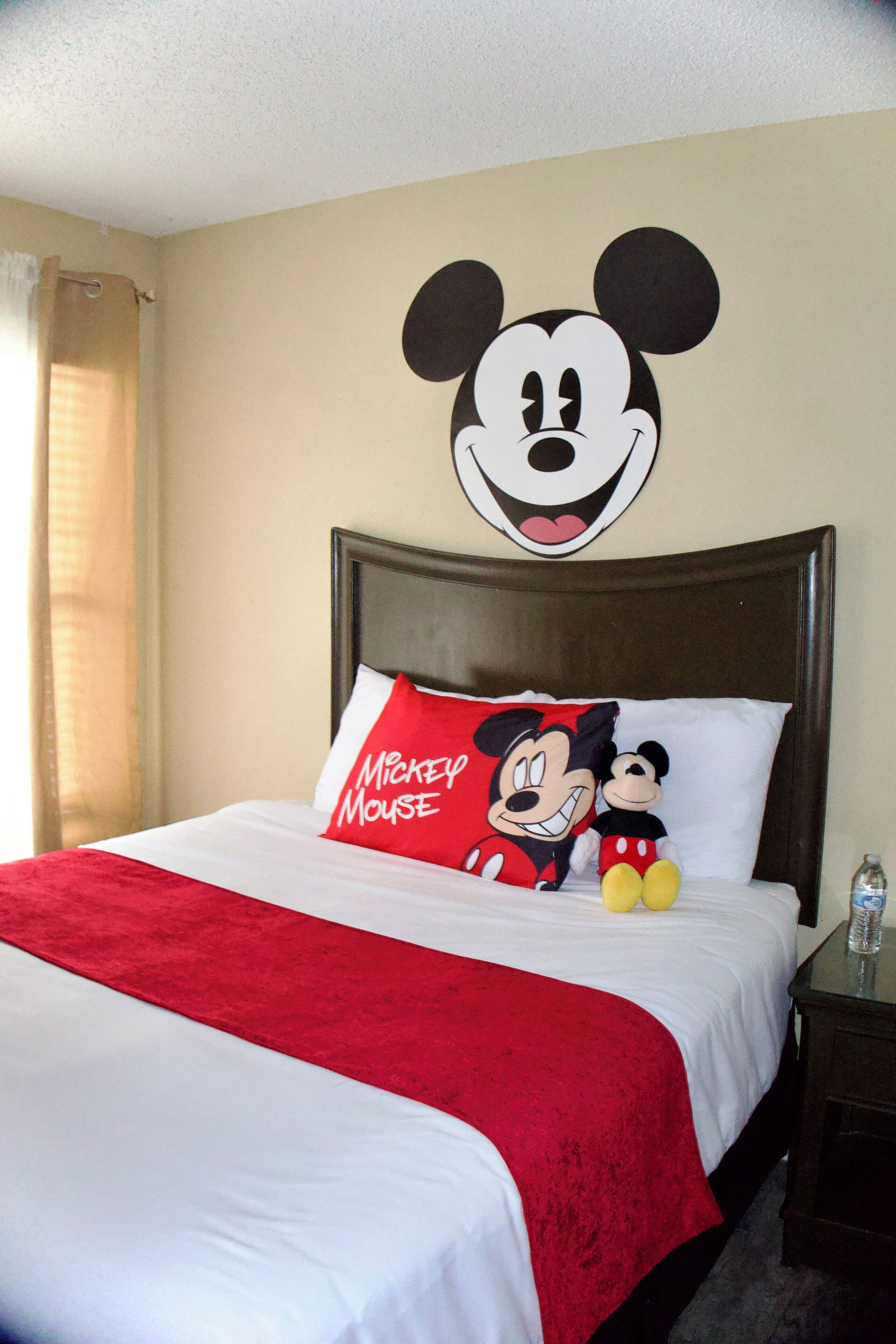 A hotel room with a large Mickey Mouse wall decal, a Mickey Mouse pillow, and a Mickey Mouse plush toy on the bed.