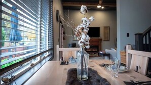 Une table à manger ornée d’un vase en verre contenant des branches de coton séchées, d’un set de table noir et moelleux, et d’une fenêtre avec des stores.