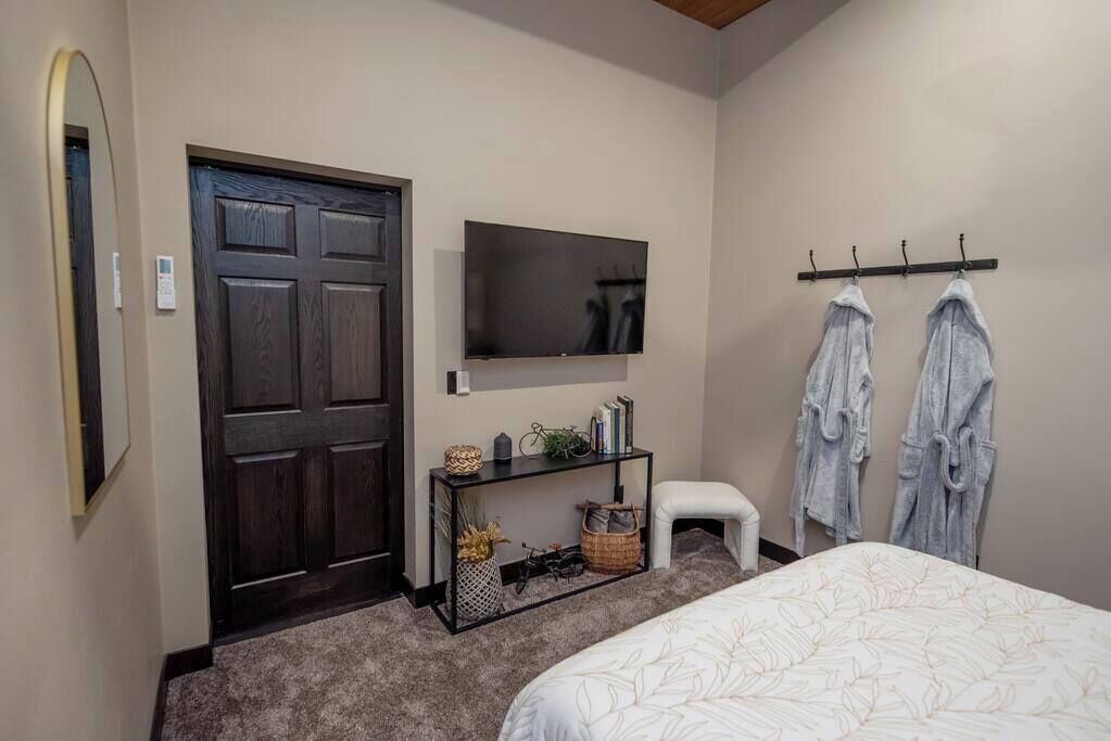 A bedroom with a bed, a TV mounted on the wall, a small table with a basket, and a door.