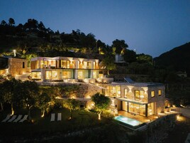 Una casa moderna de varios pisos con piscina, rodeada de vegetación y colinas.