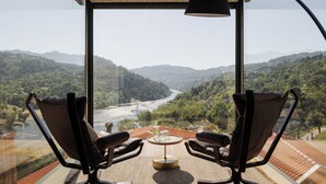 A room with a large window overlooking a valley and river, two chairs, a small table with glasses and a bottle, and a lamp. - Dajas Douro Valley – New opening (Marco de Canaveses)
