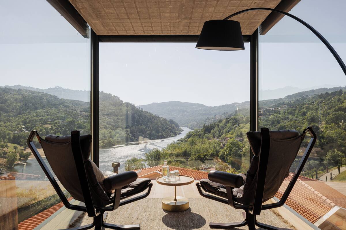A room with a large window overlooking a valley and river, two chairs, a small table with glasses and a bottle, and a lamp.