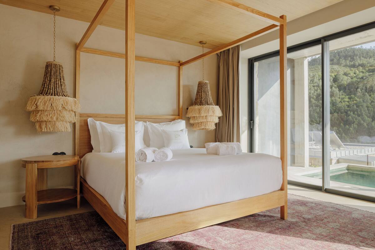A four-poster bed with white linens, a wooden nightstand, and a large window with a view of a pool and trees.