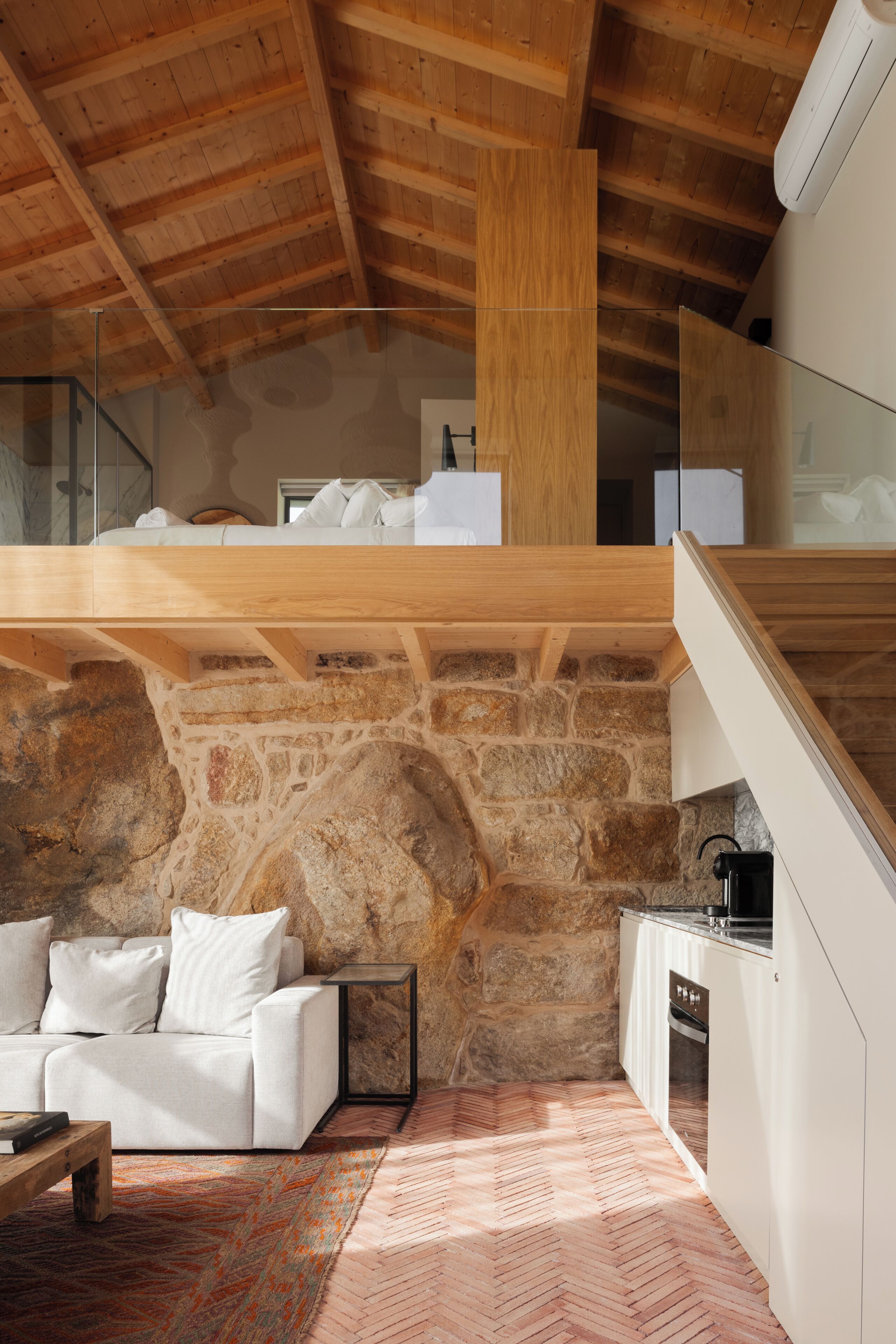 A modern interior with a stone wall, wooden ceiling beams, a white sofa, and a herringbone-patterned floor.