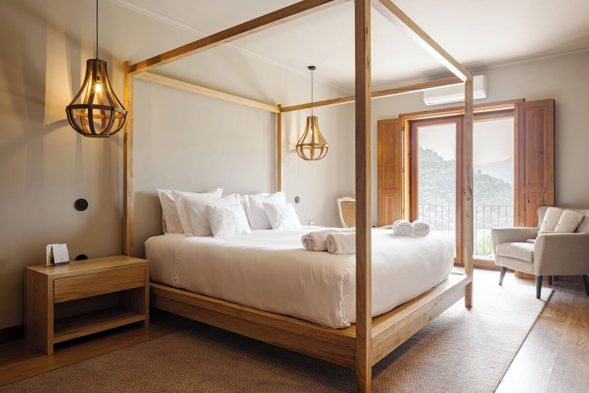 A four-poster bed with a wooden frame, a nightstand, a chair, and a view of the outdoors through a large window.