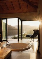 Un baño moderno con una bañera independiente, un banco de madera y vistas a un río y montañas a través de amplios ventanales.