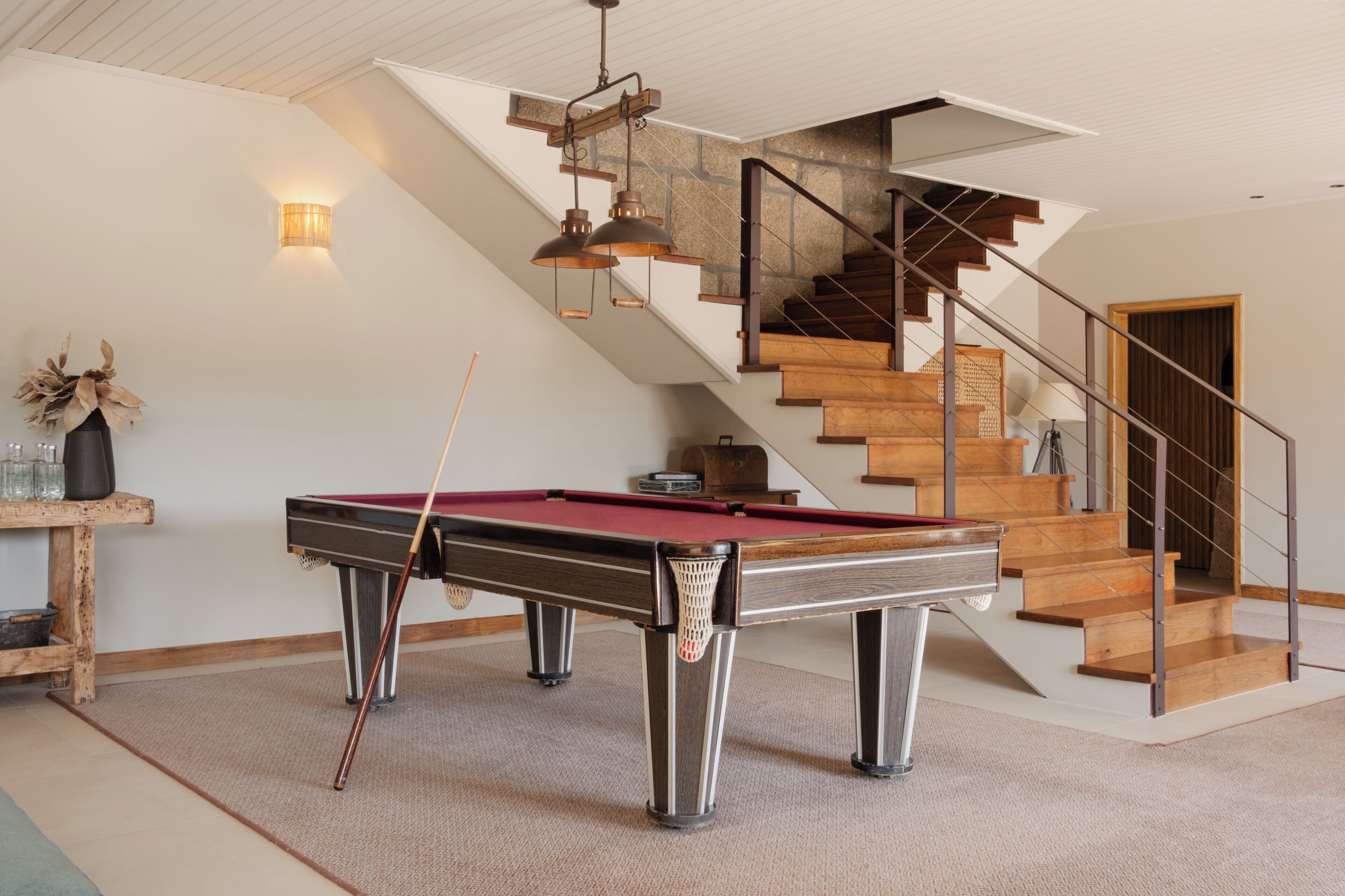A pool table with a cue stick, a wooden staircase, and a wall-mounted lamp.