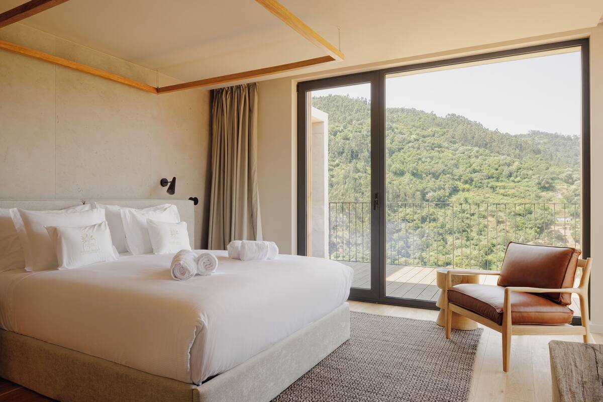 A hotel room with a large bed, a chair, a view of a forested hillside, and a balcony with a glass door.