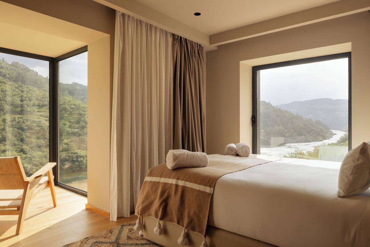 A bedroom with a large bed, a wooden chair, a view of a forested landscape through a window, and a curtain.