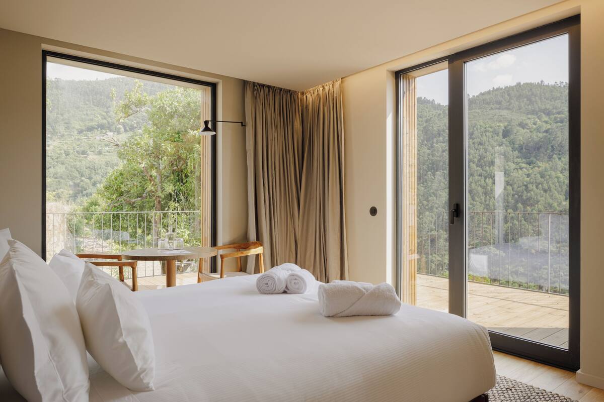 A bedroom with a large bed, a small table, and a view of a forested hillside through a sliding glass door.