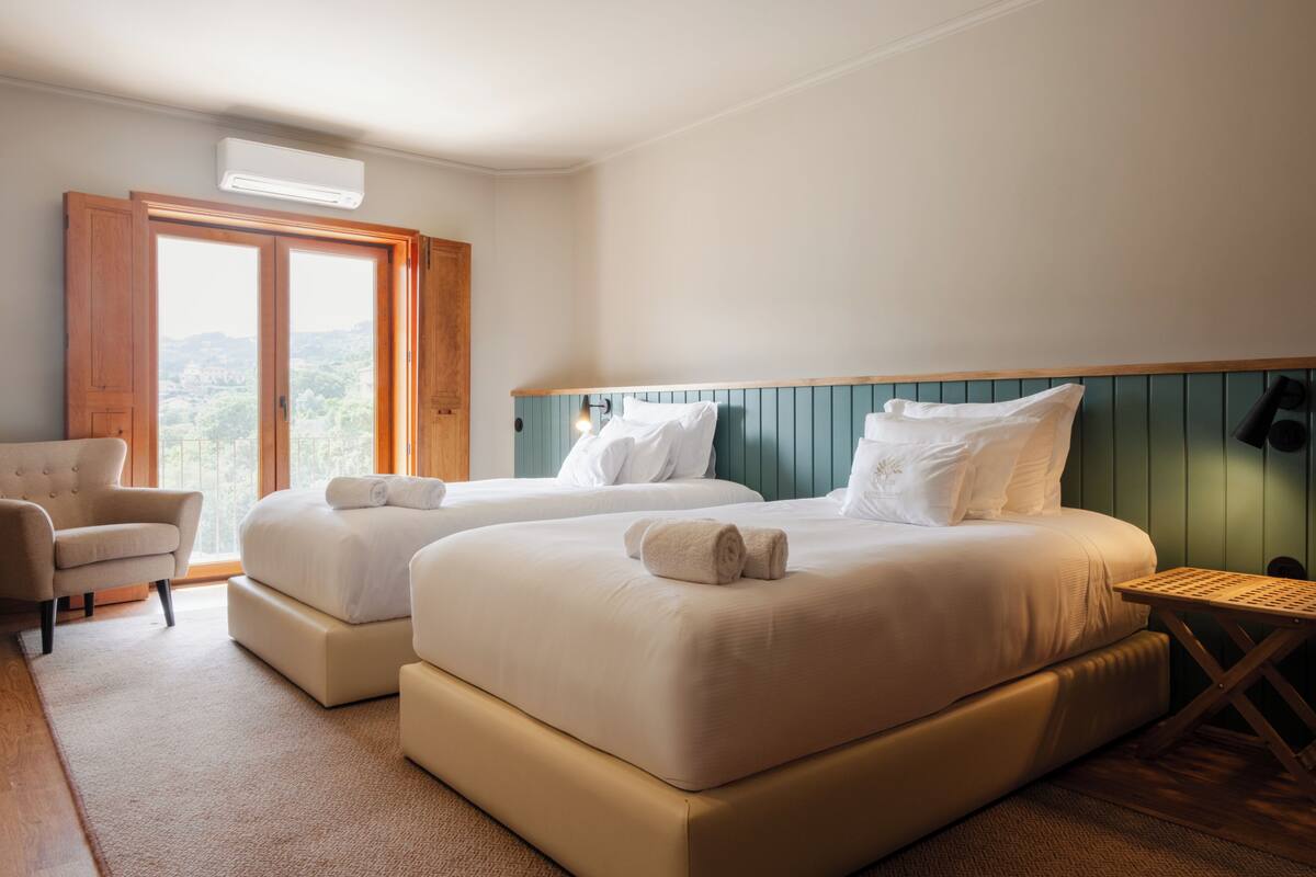A hotel room with two beds, a chair, a small table, and a view of the outdoors.