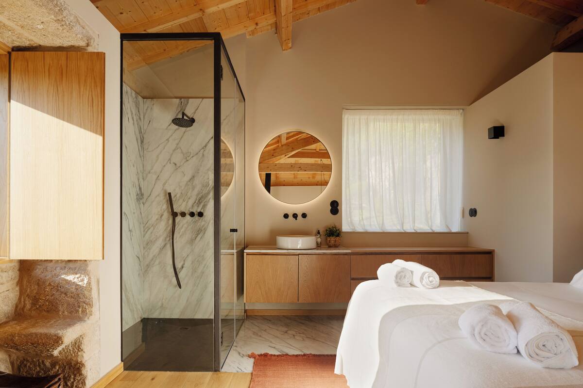 A modern bedroom with a glass-enclosed shower, wooden vanity, and a large bed with rolled towels.