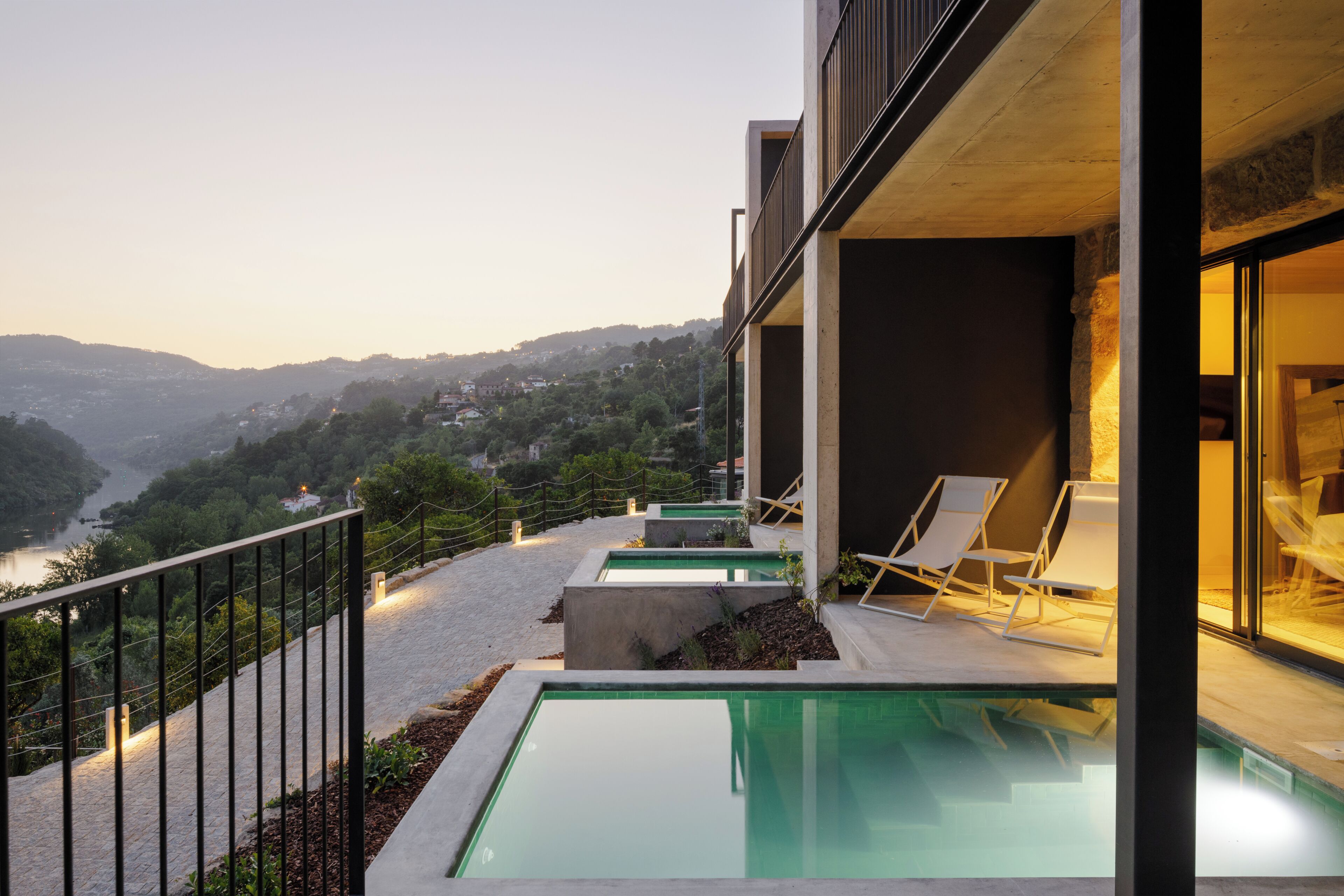 A modern building with a swimming pool, outdoor seating, and a view of a river and hills.