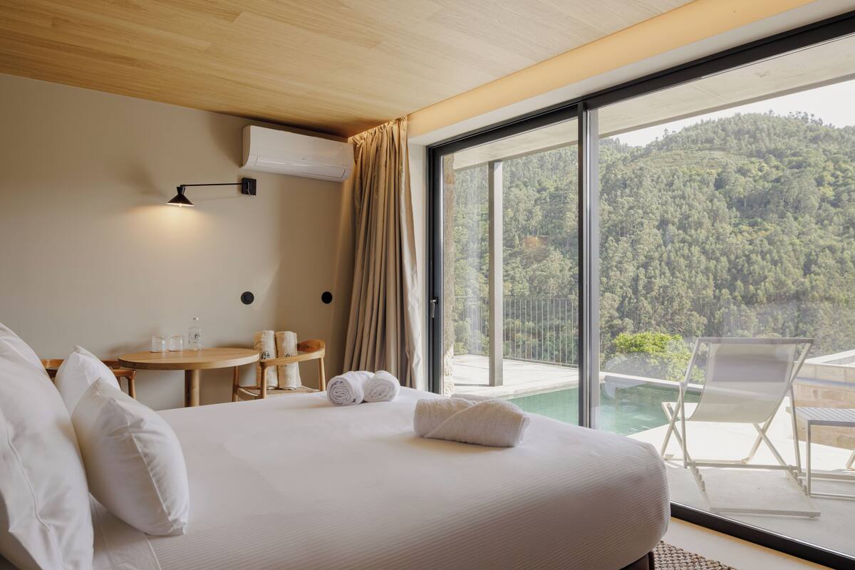 A hotel room with a bed, a small table with a chair, a window with a view of a forest, and an air conditioning unit.