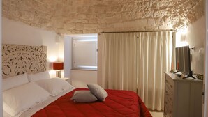 Een slaapkamer met een stenen muur, een bed met een rode deken, witte kussens, een nachtkastje met een lamp en een dressoir met een spiegel.