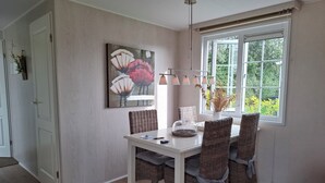 A dining area with a white table, wicker chairs, a floral painting, and a window with a view of greenery. - Pretty chalet with free WiFi on a friendly camping site (Serooskerke)