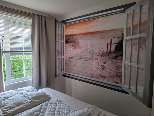 A bedroom with a bed, a window with blinds, and a wall-mounted painting of a beach scene.