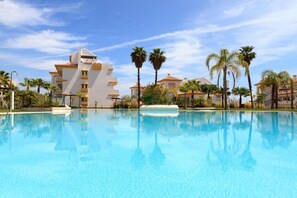 A swimming pool with a clear blue water surface, surrounded by palm trees and buildings with a Mediterranean architectural style. - CT 100 - Antes Penthouse (Mijas)