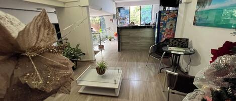 Un moderno vestíbulo de hotel con una mesa de centro de vidrio, una planta decorativa grande, una mesa pequeña con una silla y un refrigerador con imanes.