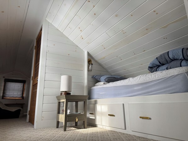 Un dormitorio con techo inclinado, una cama con ropa de cama azul, una mesita de noche con lámpara y un ventanal con persianas.