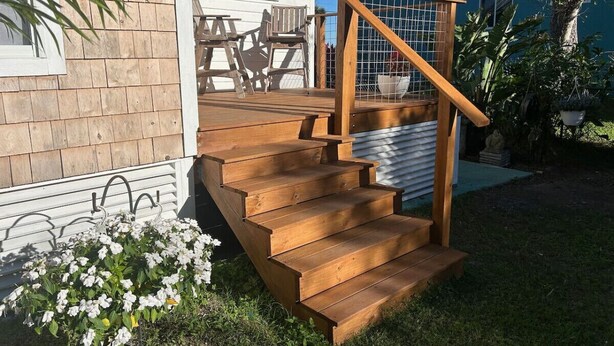 Eine Holztreppe führt zu einer Terrasse mit Geländer, umgeben von Grünpflanzen und einem Haus mit blauer Fassade.