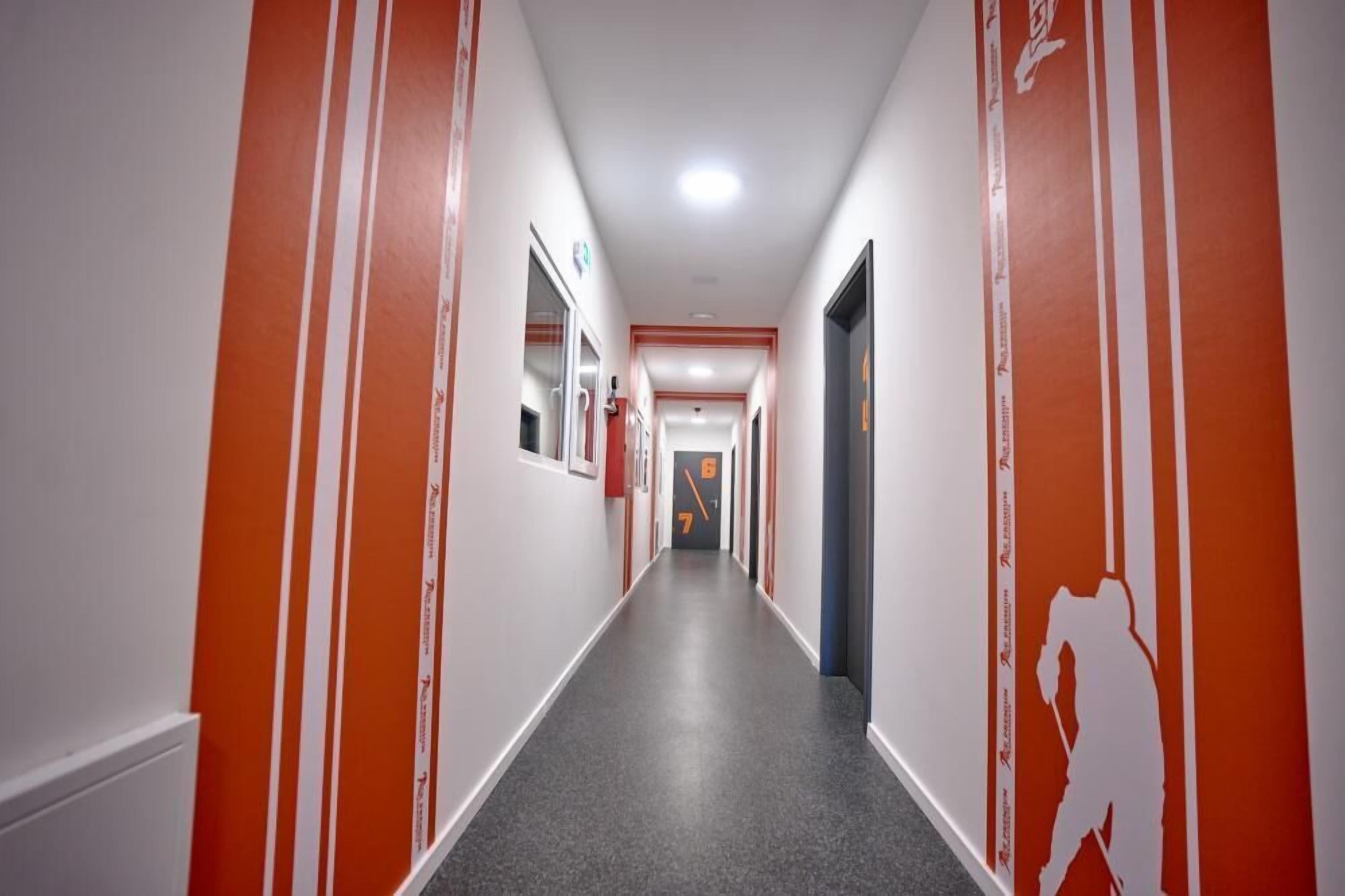 Un long couloir étroit aux murs orange et blancs, avec un tapis gris et un luminaire au plafond.