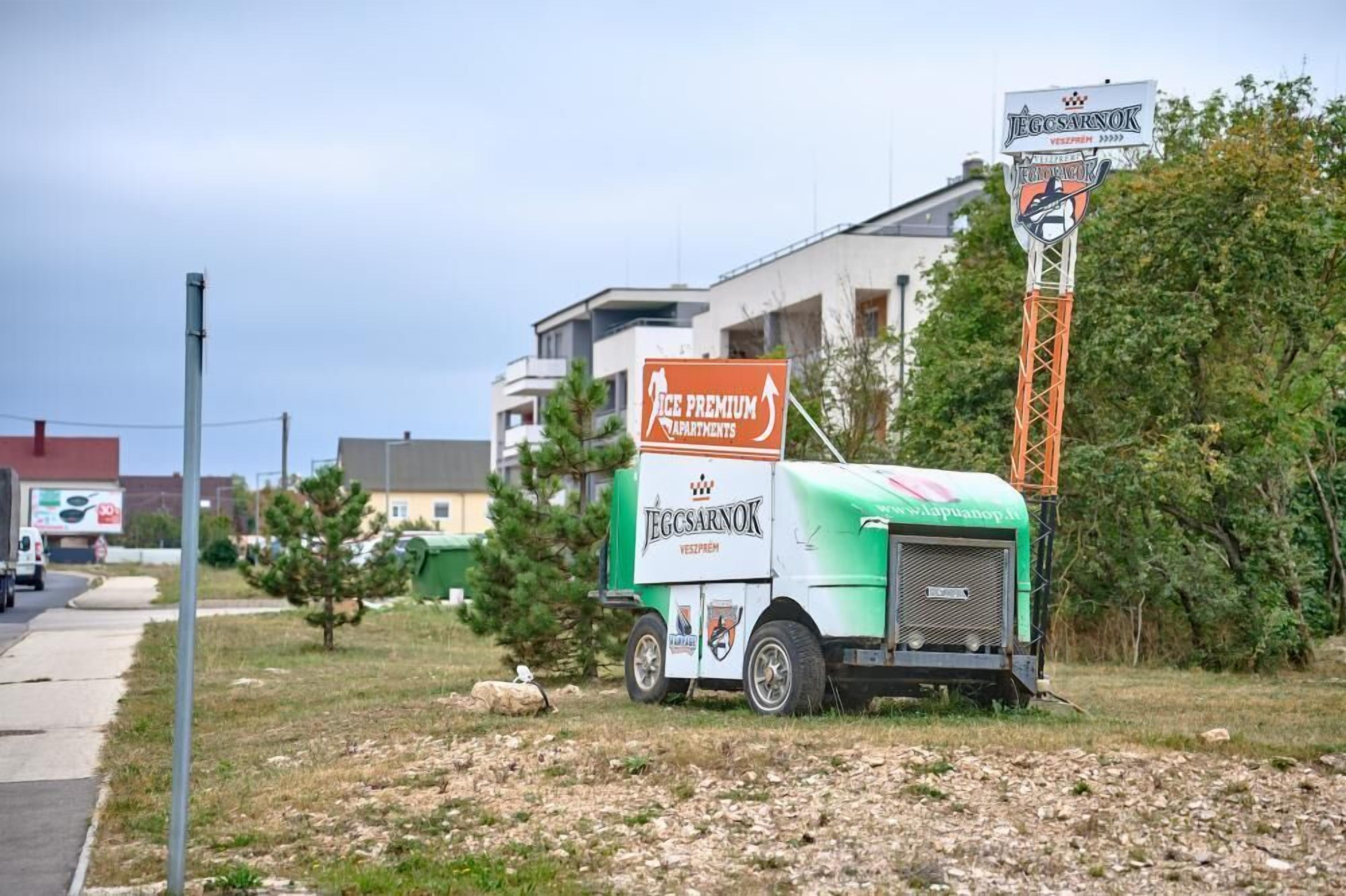 Ein grüner und weißer Umzugswagen steht auf einer Rasenfläche, im Hintergrund ein Schild für die Apartments JEGCSARNOK.