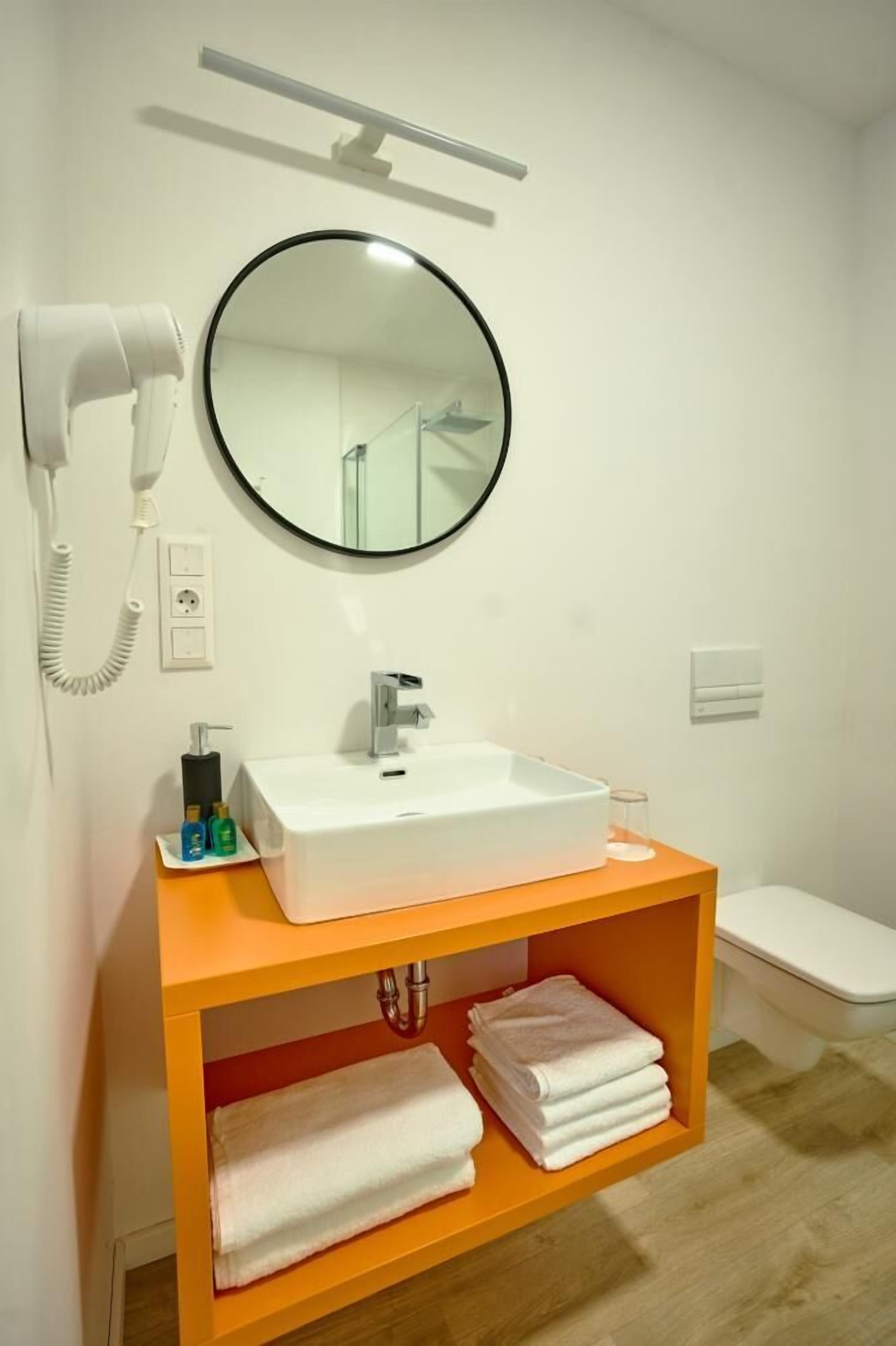 Een moderne badkamer met een witte wastafel, een ronde spiegel, een haardroger en een toilet.