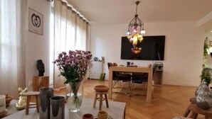 A modern living room with a wooden floor, a dining area with a table and chairs, a TV mounted on the wall, and a decorative chandelier.