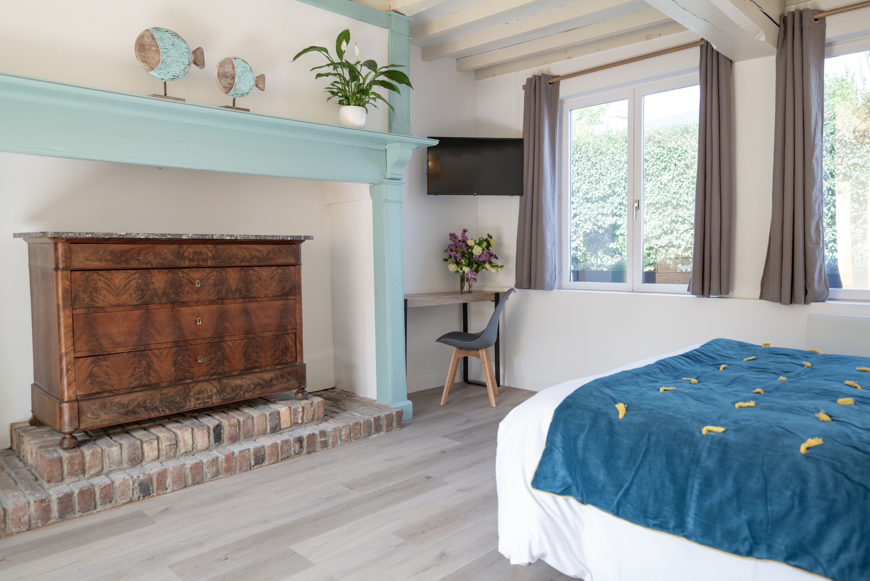 Una camera da letto con un comò in legno, una scrivania, una sedia, un letto con coperta blu, una TV montata a parete e una finestra con tende.