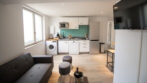 A modern kitchen with a washing machine, microwave, and refrigerator. There is a grey sofa and two stools in the living area.