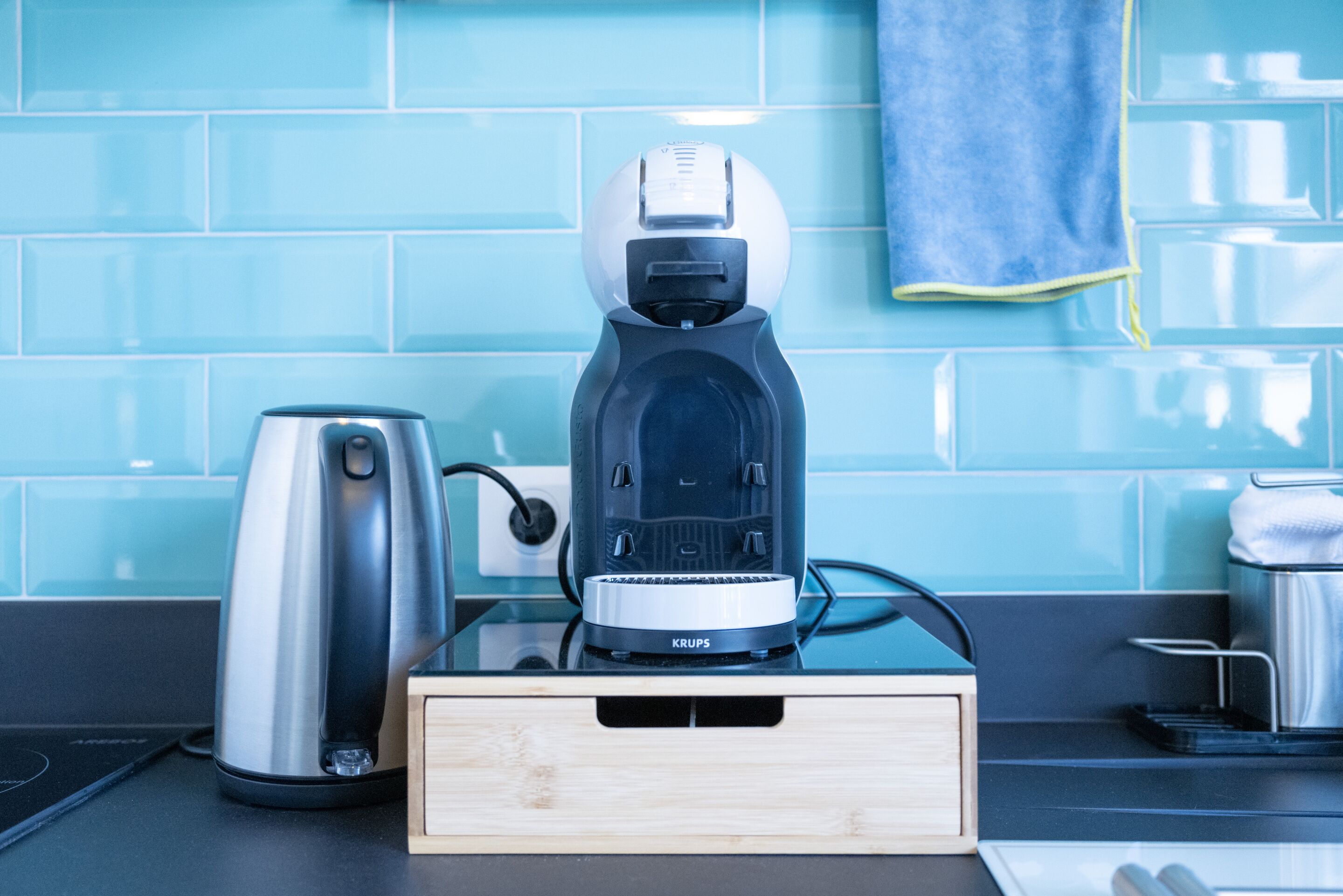Une machine à café est posée sur un comptoir en bois, avec une bouilloire à côté, un pare-feu en carreaux bleus et une serviette accrochée au mur.