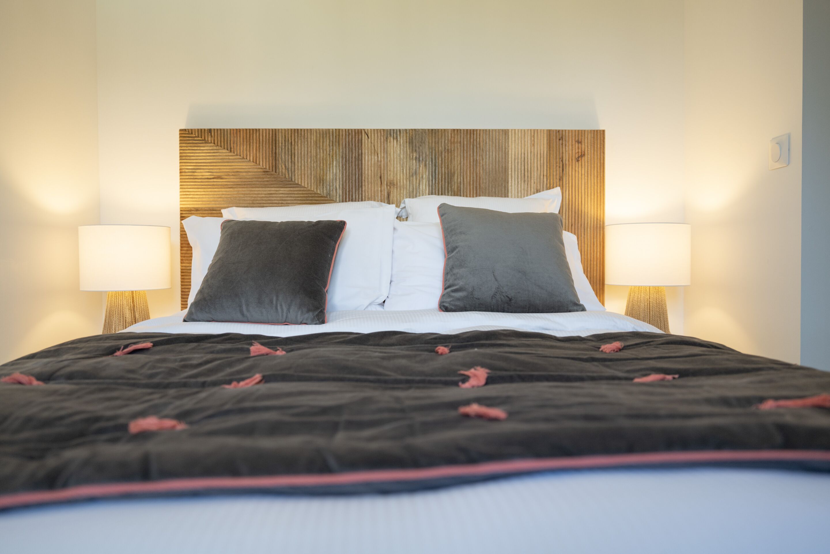 A neatly made bed with a dark blanket and two pillows, flanked by two bedside lamps.