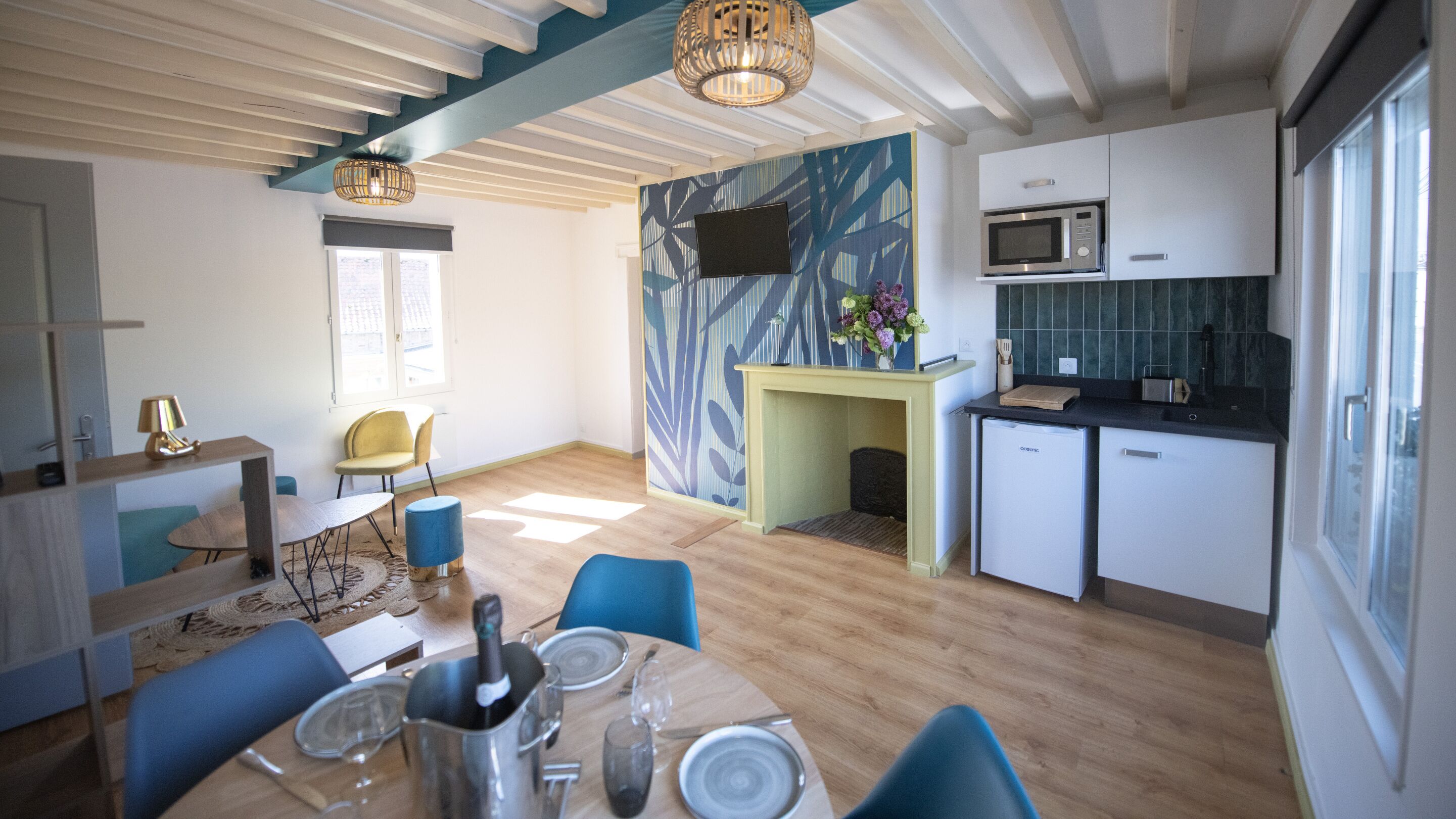 Una cocina moderna con microondas, pileta y un refrigerador pequeño. Hay una zona de comedor con una mesa dispuesta para dos personas y sillas azules. Se ve una chimenea al fondo.
