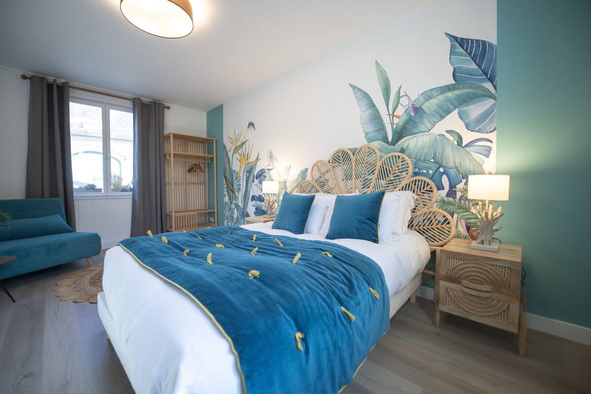Une chambre avec un grand lit, un canapé bleu, une tête de lit en bois et une fresque murale aux motifs tropicaux.