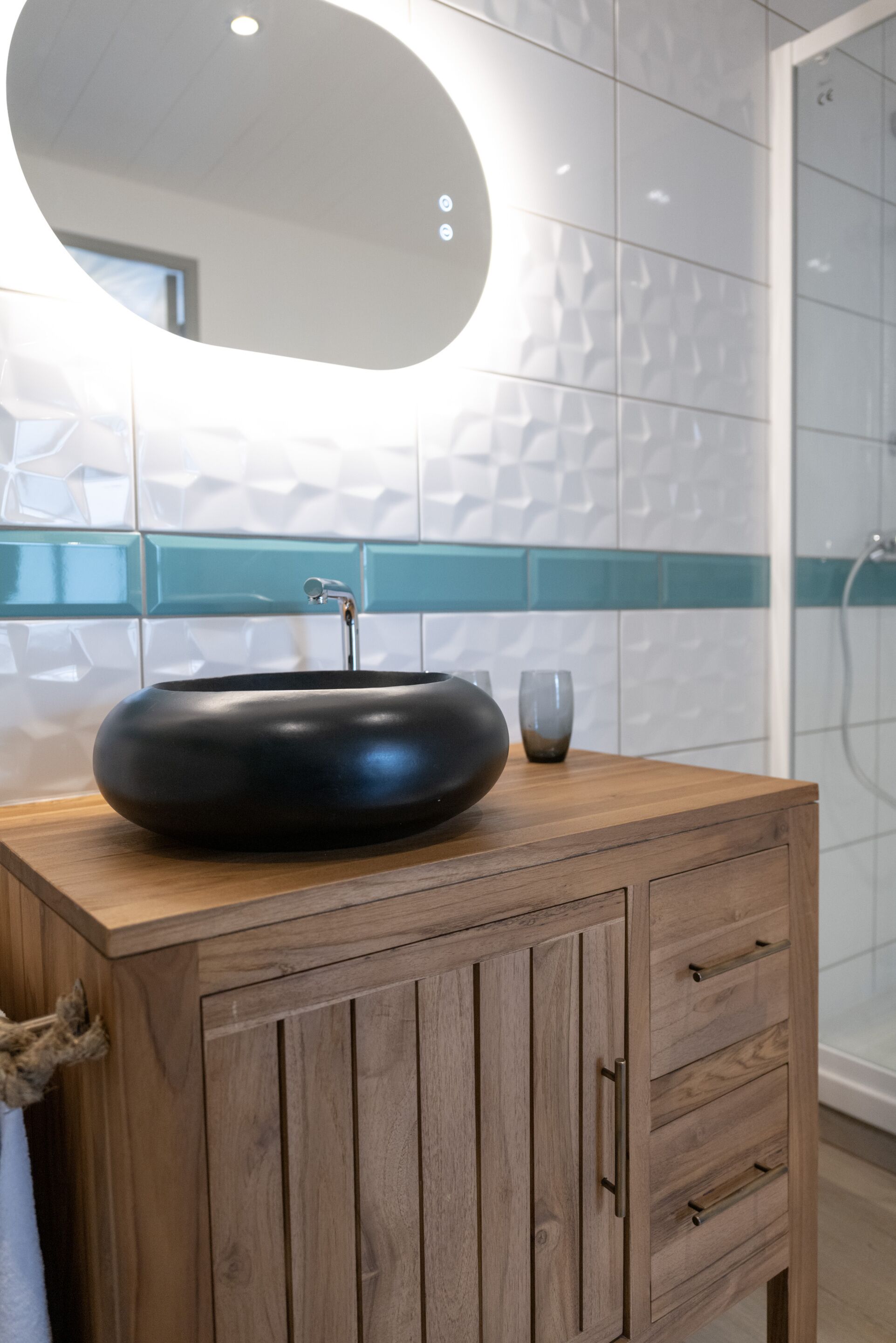 Un baño moderno con un lavamanos redondo negro, tocador de madera, azulejos verde azulado y blancos, y una cabina de ducha de vidrio.