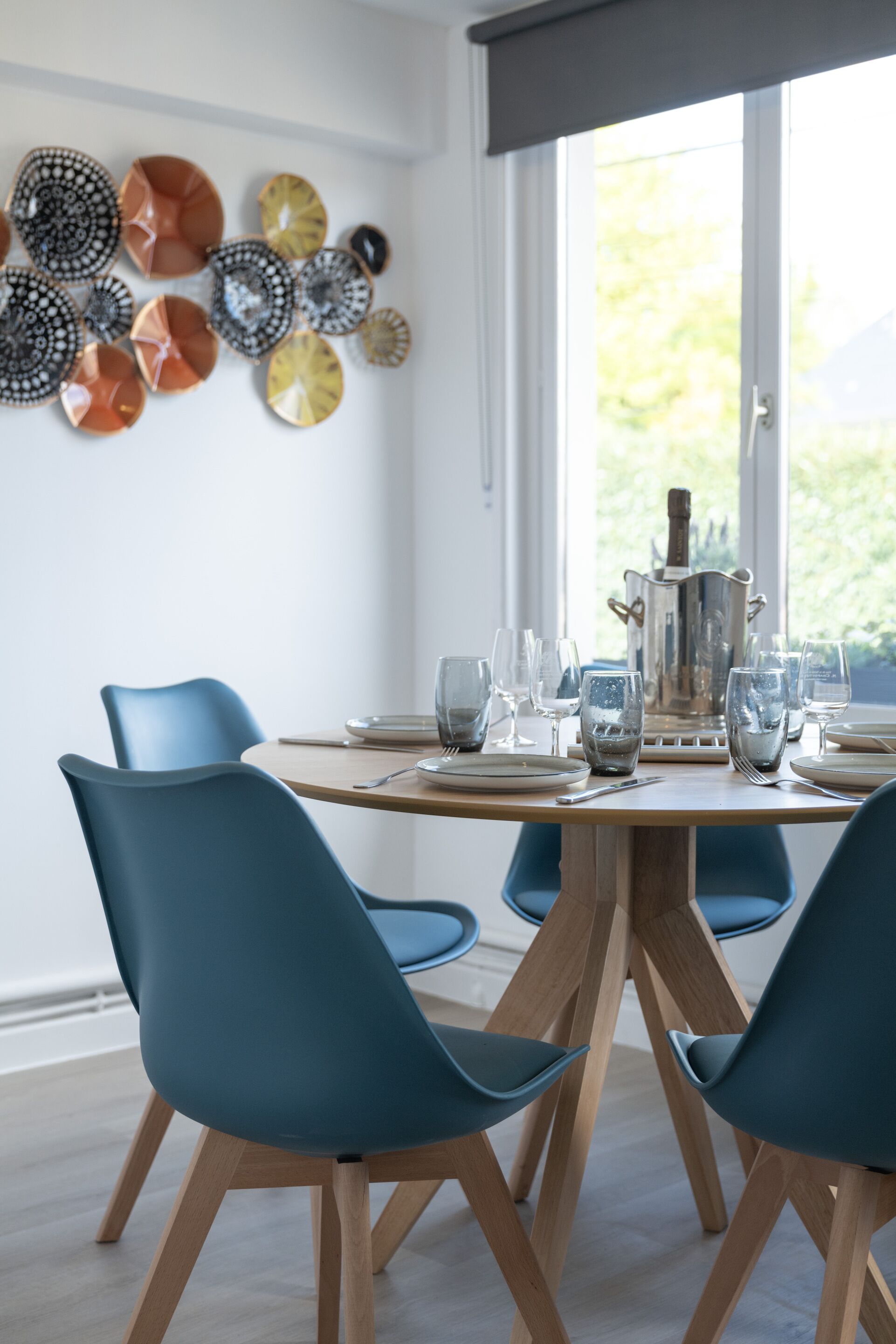 Un coin repas avec une table ronde, des chaises bleues et un mur décoratif orné d’assiettes.