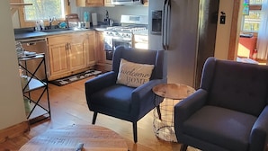 Een knus interieur van een blokhut met een keuken, twee fauteuils, een ronde salontafel en een vloerkleed.