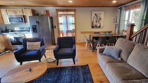 Een gezellige woonkamer met een bank, fauteuil en salontafel. In de achtergrond is een keuken met moderne apparatuur zichtbaar.