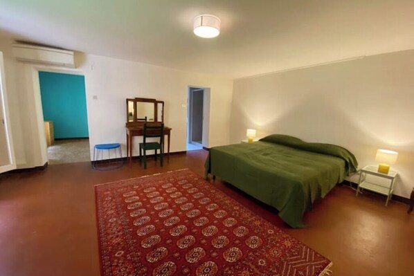 Un dormitorio con cama, un escritorio con silla, una mesita de noche con lámpara y una alfombra estampada.
