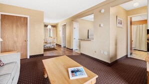 Habitación de hotel con puerta de madera, zona de comedor con mesa y sillas, kitchenette con refrigerador y baño visible a través de una puerta abierta.