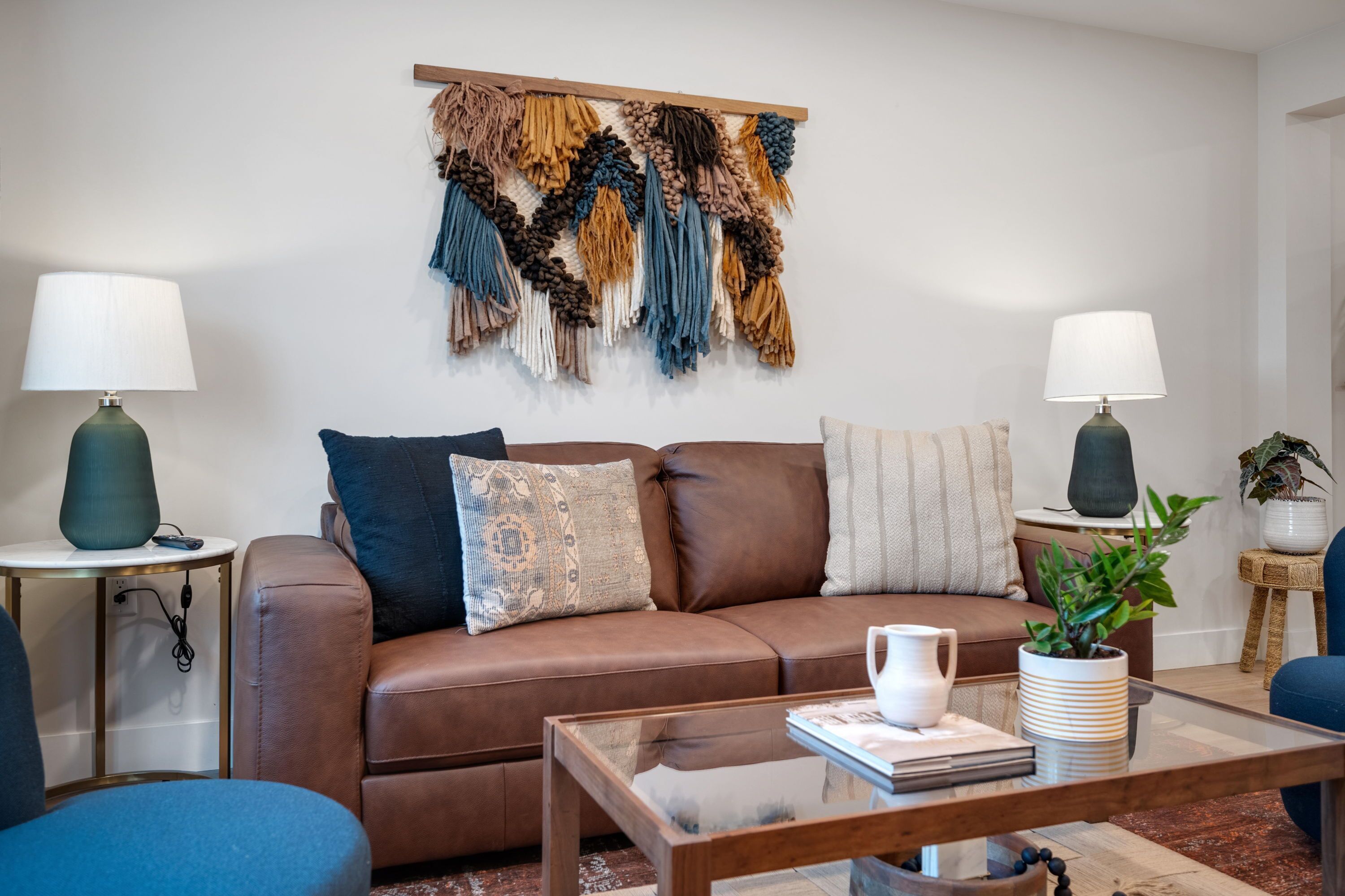 Uma sala de estar moderna com um sofá de couro marrom, duas mesas laterais com abajures, uma mesa de centro de vidro e um painel de macramé na parede.
