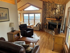 Un salon chaleureux avec une cheminée en pierre, des meubles en bois et de grandes fenêtres donnant sur l’extérieur.