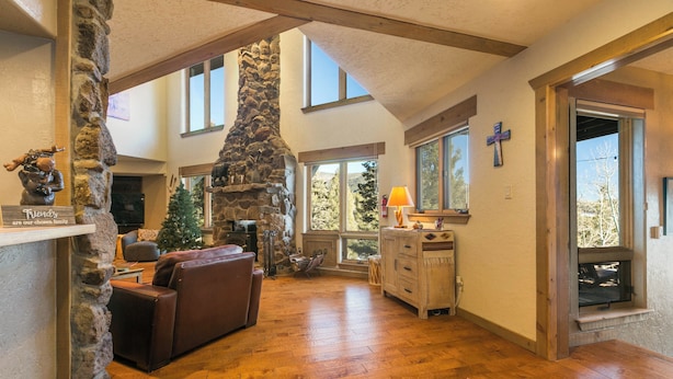 A spacious living room with a stone fireplace, wooden beams, and large windows offering views of the outdoors. - Elevated Home w/Hot Tub, Beautiful Views, Firepit, 3 King Bedrooms, Bunkroom (Angel Fire)