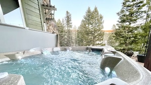 A hot tub with a stone surround, set against a backdrop of trees and a building. - Elevated Home w/Hot Tub, Beautiful Views, Firepit, 3 King Bedrooms, Bunkroom (Angel Fire)