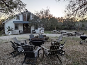 Une maison avec une cour en gravier, un espace extérieur aménagé avec des fauteuils Adirondack, un foyer et des tables de pique-nique.