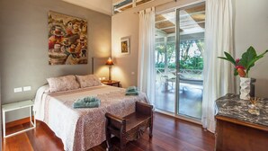 Une chambre à coucher avec un grand lit, un banc en bois, une table de chevet, une lampe, un tableau et une vue sur l’extérieur grâce à une porte-fenêtre coulissante.