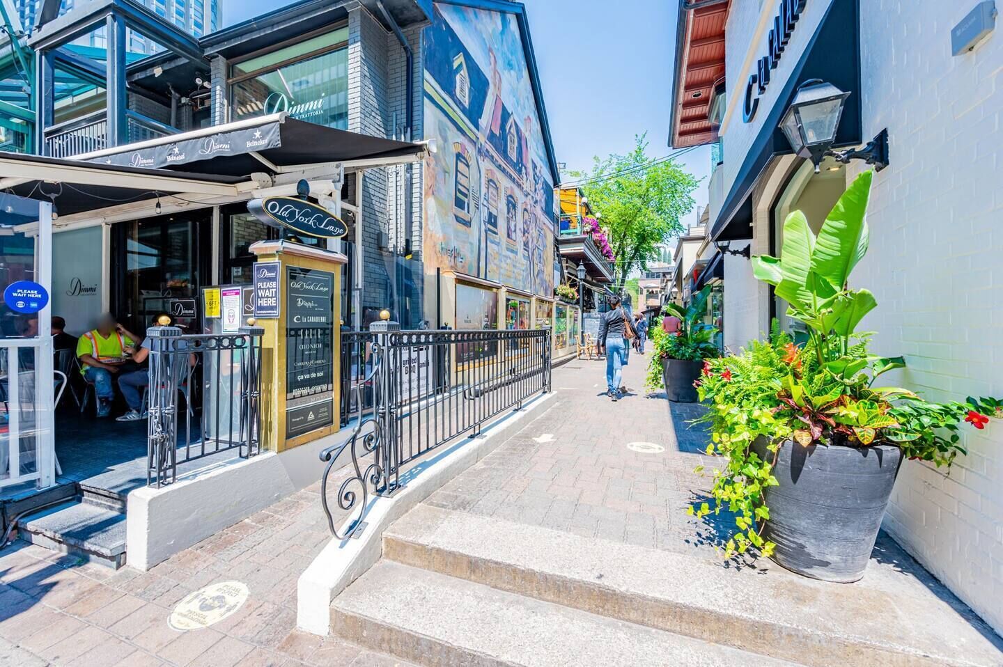 A narrow street with buildings on either side, a café with outdoor seating, and a large planter with tropical plants.