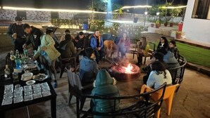 An outdoor dining area with a fire pit, tables, and chairs, surrounded by decorative lights and greenery. - Camp Aquaforest  (Rishikesh)